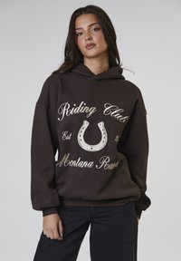 Jonge vrouw met lang donker haar, gekleed in een donkerbruine hoodie met de tekst "Riding Club Montana Ranch" en een hoefijzerafbeelding.