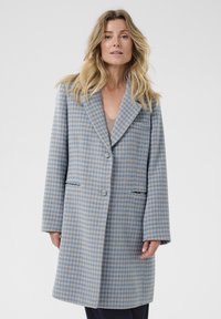 Cappotto azzurro chiaro con motivo a scacchi, caratterizzato da un ampio rever, due tasche frontali e una vestibilità rilassata. Tessuto liscio con una texture morbida.