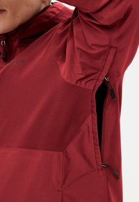 Veste pull rouge en tissu texturé, avec poche zippée et design à panneaux contrastants. Légère et conçue pour le confort.