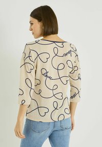 Top beige de manga 3/4 con un patrón de corazón azul marino y cursiva, con cuello redondo y ajuste holgado. Tela texturizada con aberturas laterales.