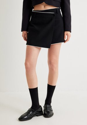 Zwarte wikkelrok met witte bies, voorzien van een knoopsluiting en zijzak. Gestyled met zwarte geribde sokken en glanzende loafers.