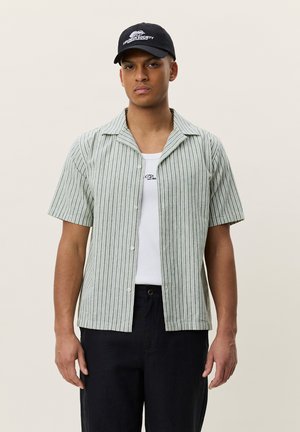 Mand iført sort kasket, hvid ribbet tanktop, åben grøn- og hvidstribet kortærmet skjorte og sorte bukser, stående mod en ensfarvet baggrund.