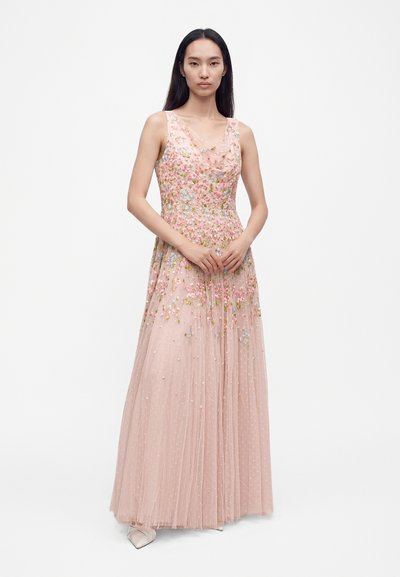 Femme aux longs cheveux foncés portant une robe maxi rose clair sans manches brodée de fleurs, debout devant un fond blanc uni.