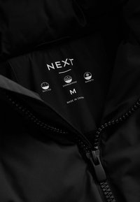 Čierna prešívaná bunda zvnútra s logom značky "NEXT." Obsahuje ikony pre odolnosť voči dažďu, teplo, veľkosť "M" a vyrobené v Číne.