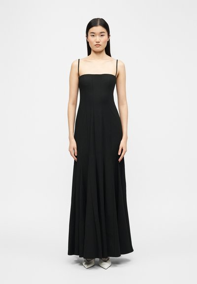 Femme aux cheveux longs et raides portant une robe noire sans manches longue jusqu'au sol et des chaussures blanches pointues, debout devant un fond uni.