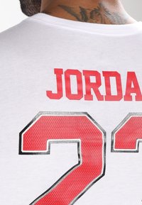 Vit bomull t-shirt med röd text "JORDAN" och siffran "23" i en djärv, perforerad design med svart kontur.