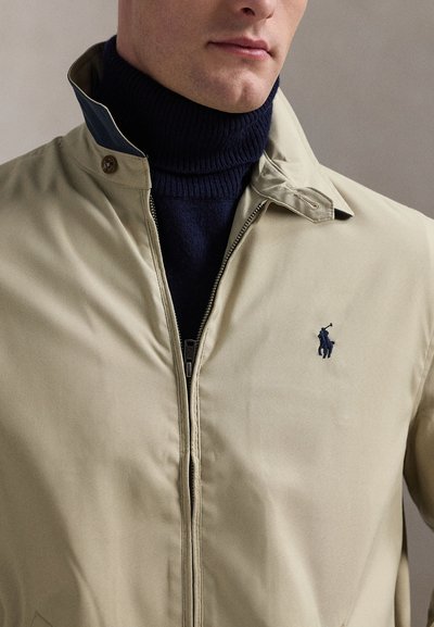 Mand iført beige lynlåstrøje over en navyblå rullekrave, med et lille mørkt broderet polo-spillerlogo på jakken.