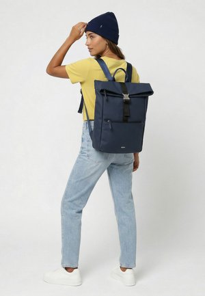 Femme portant un bonnet bleu marine, un t-shirt jaune, un jean bleu clair, des baskets blanches, et un sac à dos roll-top bleu marine avec des détails de boucle et de fermeture éclair.