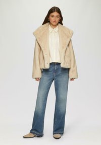 Beige faux fur jas met een brede kraag, gecombineerd met een wit gestreept shirt en uitlopende blauwe spijkerbroek. Opvallende details zijn de zachte textuur.