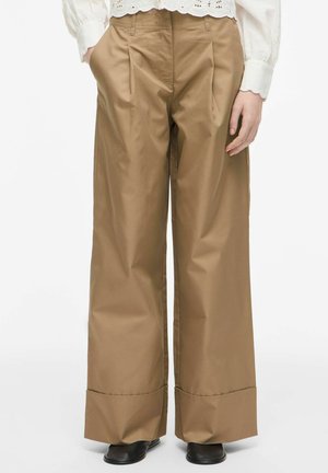 Pantalon beige à jambes larges avec plis devant, ourlets retroussés, poches latérales, associé à des chaussures noires et un haut blanc à manches longues.