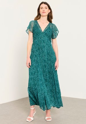 Femme portant une robe maxi à fleurs bleu sarcelle avec un col en V, des manches volantes transparentes et des sandales blanches à talons, debout contre un mur uni.