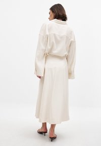 Lång beige klänning med en något lös passform, som har en krage på baksidan och långa ärmar. Slät textur, som når till mitten av vaderna, i kombination med låga klackar.