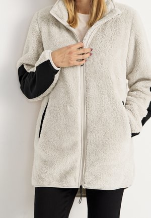 Chaqueta de forro polar color crema con mangas de nailon negro, bolsillos frontales y cremallera completa. Textura suave y ajuste relajado, diseño de largo medio.