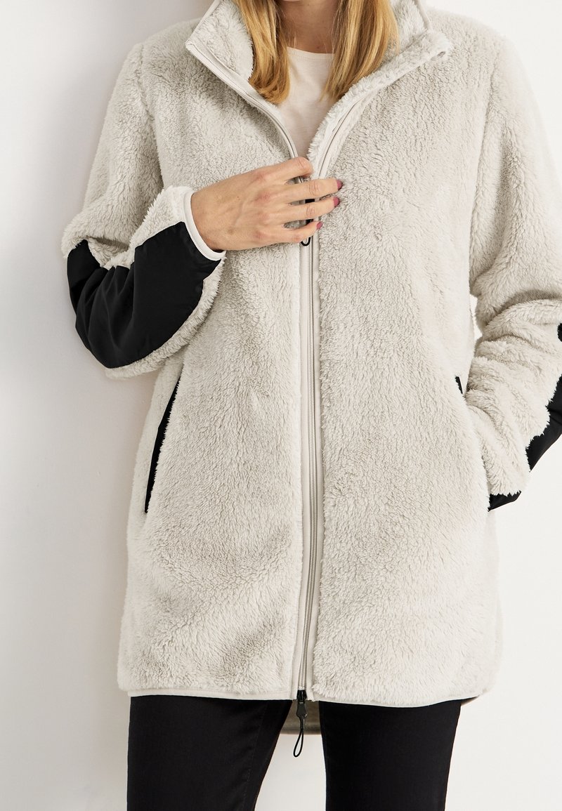 Cremefarbige Fleecejacke mit schwarzen Nylonärmeln, Fronttaschen und einem durchgehenden Reißverschluss. Weicher Stoff und lässige Passform, mittellanges Design.