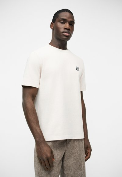 Νεαρός άνδρας που φοράει ένα απλό κοντομάνικο μπλουζάκι σε εκρού χρώμα με μικρό λογότυπο λύκου στο στήθος και υφασμένα μπεζ παντελόνια.