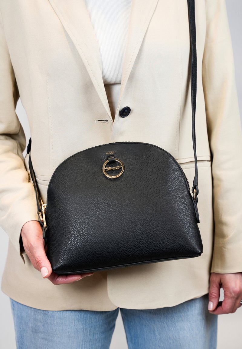 Sac à main en cuir noir avec un dessus arrondi, surface texturée, détail de quincaillerie circulaire dorée et une bandoulière amovible.