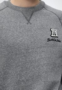 Gros plan d'un pull gris à col rond avec un tissu texturé et un logo brodé représentant une bobine avec le texte "Scotch & Soda" sur la poitrine.