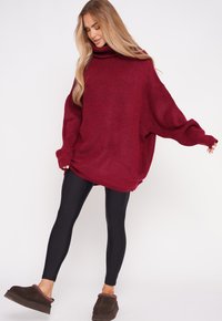 Burgundowy, oversize sweter z wysokim kominem, teksturowana tkanina, długie rękawy, w połączeniu z czarnymi legginsami i ciemnobrązowymi butami na slip-on.