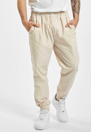 Beige, lightweight jogger pants with elastic waistband, drawstring, and side pockets. Cuffs are elasticated. Paired with white sneakers.