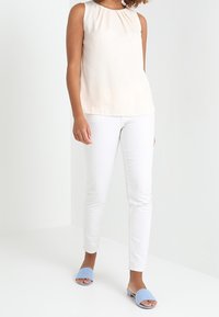 Top sin mangas de color rosa claro, jeans ajustados blancos y sandalias deslizantes azules. Tejido suave con detalle de escote fruncido, silueta limpia.