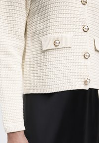 Kremfarvet strikket cardigan med tekstureret mønster, guldbesatte knapper med perler, klaplommer, båret over en sort nederdel eller kjole.