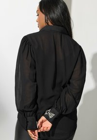 Blusa negra transparente con mangas abullonadas, un cuello clásico y puños decorados con joyas y adornos florales.