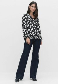 Blus med svart och vit mönster har en V-ringning, veckade puffärmar och en avslappnad passform, ihop med mörka utsvängda jeans och svarta skor.