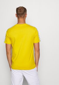 Camiseta de algodón amarilla con mangas cortas, que presenta un patrón en contraste a lo largo de la costura del hombro y un diseño clásico de cuello redondo.