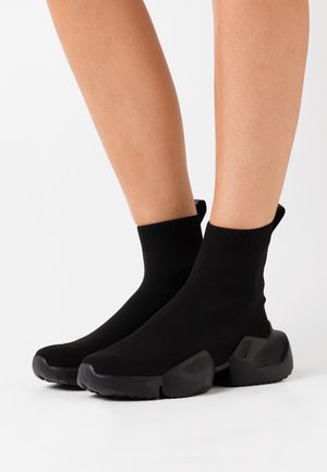 Benen met zwarte sokachtige sneakers met dikke, gestructureerde zolen op een witte achtergrond.
