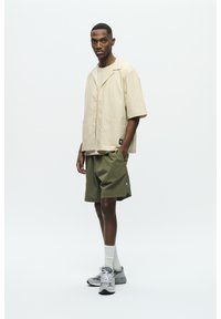 Helles beige Kurzarmhemd, leichtes Material, zwei frontale Taschen. Olivgrüne Shorts, lockere Passform. Graue Sneaker mit weißen Akzenten.