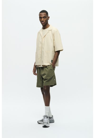 Chemise courte à manches courtes beige clair, en tissu léger, avec deux poches avant. Shorts vert olive, coupe ample. Baskets grises avec des accents blancs.