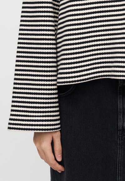 Ριγέ πουλόβερ με μαύρες και κρεμ οριζόντιες ρίγες, υφασμάτινη υφή και φαρδιά μανίκια, συνδυασμένο με σκούρο τζιν παντελόνι.