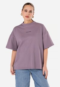 Lavendel värviline oversize t-särk, mis on valmistatud puuvillast, ümar kaelus, lühikesed varrukad ja esiküljel tumedamas variandis trükitud logo.
