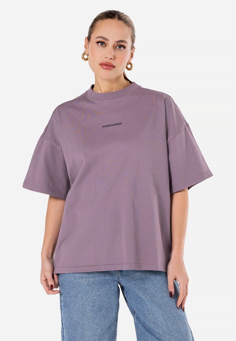 Lavendel värviline oversize t-särk, mis on valmistatud puuvillast, ümar kaelus, lühikesed varrukad ja esiküljel tumedamas variandis trükitud logo.