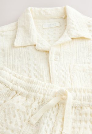 Komplet bombažnega domačega oblačila v barvi slonove kosti, ki vključuje srajco s ovratnikom in zapiranjem na gumbe ter teksturirane hlače z elastičnim pasom in vrvico za zavezovanje.