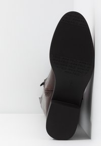 Semelle de bottine marron foncé montrant un talon et l'avant du pied texturés avec un motif répété "ANNA FIELD" et des marquages de taille.