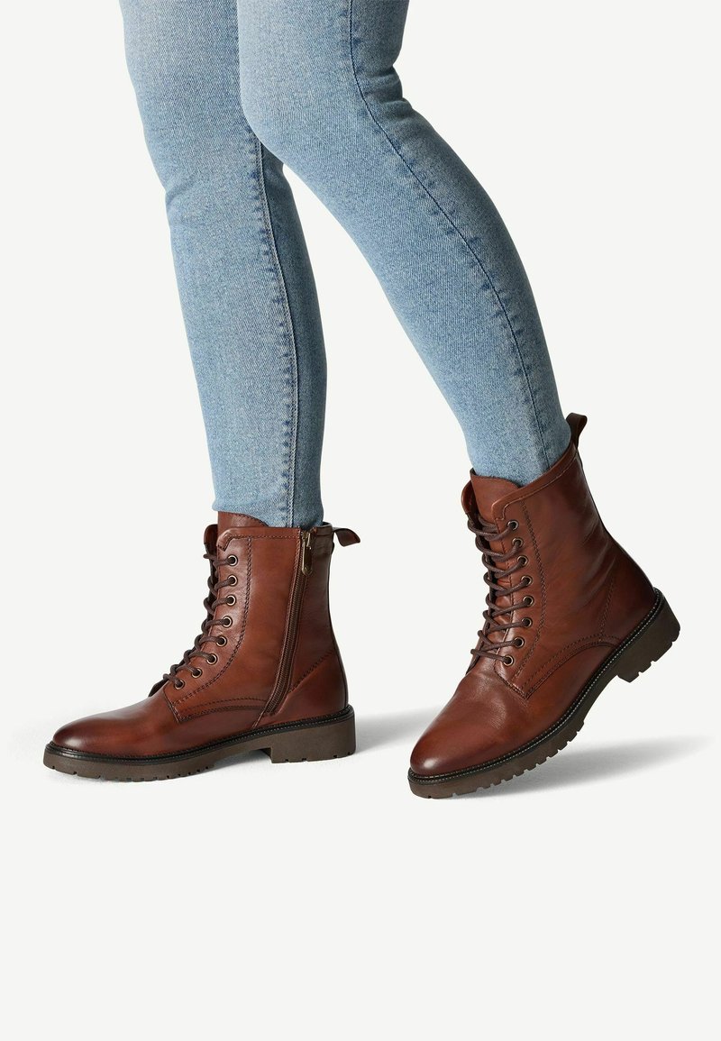 Stivali alla caviglia in pelle marrone con una finitura liscia, lacci anteriori, cerniera laterale e suola in gomma texture, abbinati a jeans di denim chiaro.