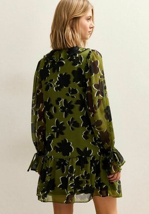 Robe fleurie vert olive, ornée de motifs floraux noirs et blancs, manches longues avec poignets froncés, et ourlet à volants.