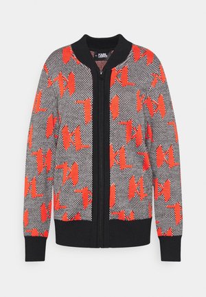 Cardigan tricoté à fermeture éclair complète avec col, poignets et ourlet noirs, présentant un motif noir et blanc surmonté de grandes formes abstraites rouges.