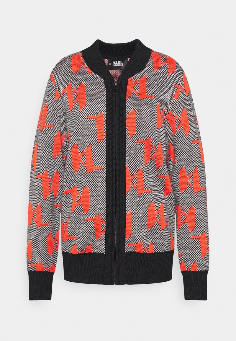 Cardigan tricoté à fermeture éclair complète avec col, poignets et ourlet noirs, présentant un motif noir et blanc surmonté de grandes formes abstraites rouges.