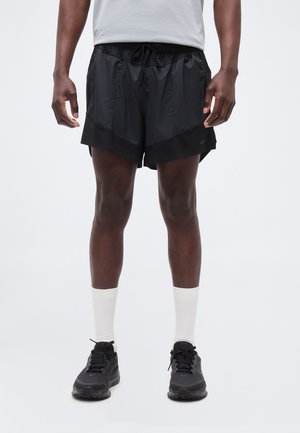 Homme portant un short de sport noir, des chaussettes blanches et des chaussures de course noires, debout, les bras détendus le long du corps.