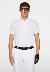 Vit kortärmad pikétröja med krage, svart bälte, vita byxor och svart golfhandske. Modellen har stora, tonade solglasögon.