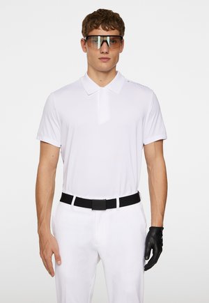 White short-sleeve polo shirt with a collar, black belt, white pants, and black golf glove. The model wears large, tinted sunglasses.