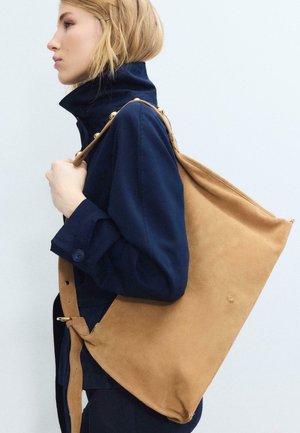 Plavolaska ženska v mornarsko modri jakni nosi veliko rjavo zamšeno torbo čez ramo z odstranljivim pasom na enobarvnem ozadju.