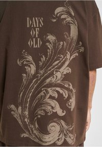 Brun bomulds t-shirt med et stort lys beige svirvelmønster på ryggen, med teksten "DAYS OF OLD" i stiliseret skrifttype.
