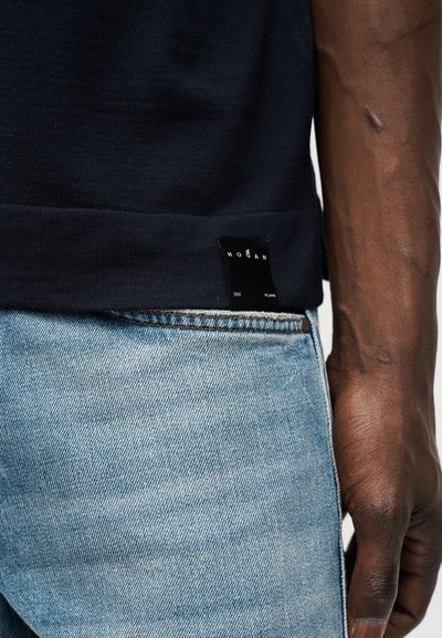 Κλειστό πλάνο ενός ατόμου που φοράει μαύρο πουκάμισο με ετικέτα Hogan και ανοιχτού χρώματος τζιν, δείχνοντας το δεξί του χέρι και βραχίονα.