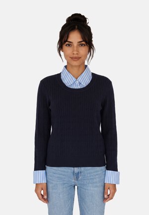 Donna con capelli scuri raccolti in uno chignon che indossa un maglione a trecce blu navy sopra una camicia a righe blu con colletto e jeans azzurri su uno sfondo semplice.