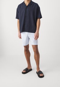 Camisa de botones de manga corta en navy, pantalones cortos a rayas azul claro y sandalias de diapositivas negras, que muestran un ajuste relajado y un estilo informal.