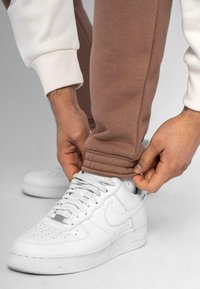Hvide Nike Air Force 1 sneakers med grå snørebånd, båret med brune sweatpants med en snæver afslutning. Hænder justerer bukseafslutningen over skoen.