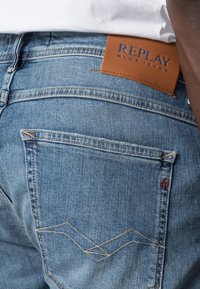 Jeans de mezclilla azules con un parche de cuero marrón que muestra "REPLAY BLUE JEANS". Presenta una superficie texturizada y un diseño distintivo de costura en el bolsillo trasero.